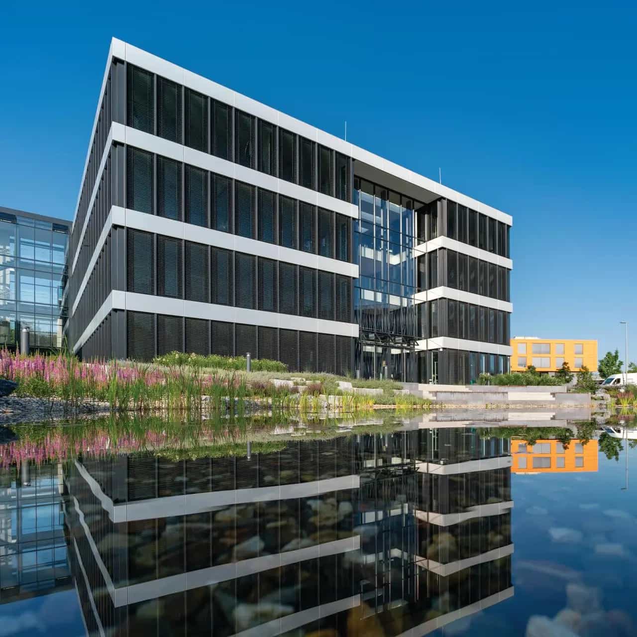 Kantoorgebouw van het hoofdkantoor van Goldbeck in Bielefeld aan een vijver, omringd door bloeiende planten onder een blauwe hemel.